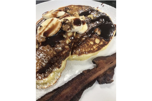 A close up photo of pancakes with bananas and chocolate syrup on them and a crispy piece of bacon on the side.